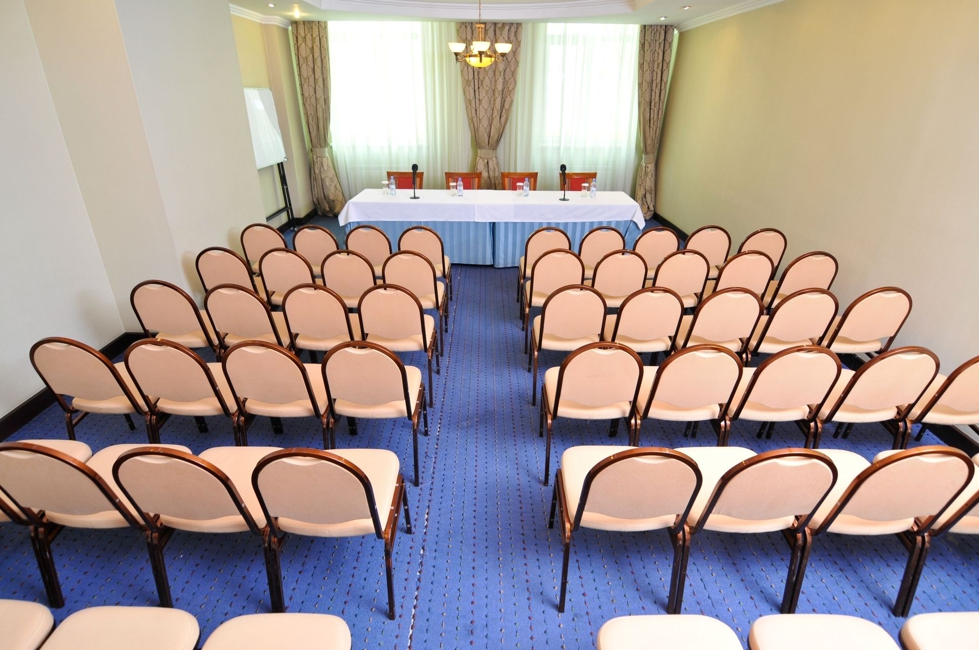 A conference room with rows of chairs facing a table with microphones, a whiteboard, and a chandelier.