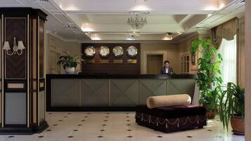 Un vestíbulo de hotel con una recepción, varios relojes, una araña de cristal y una planta grande en maceta.
