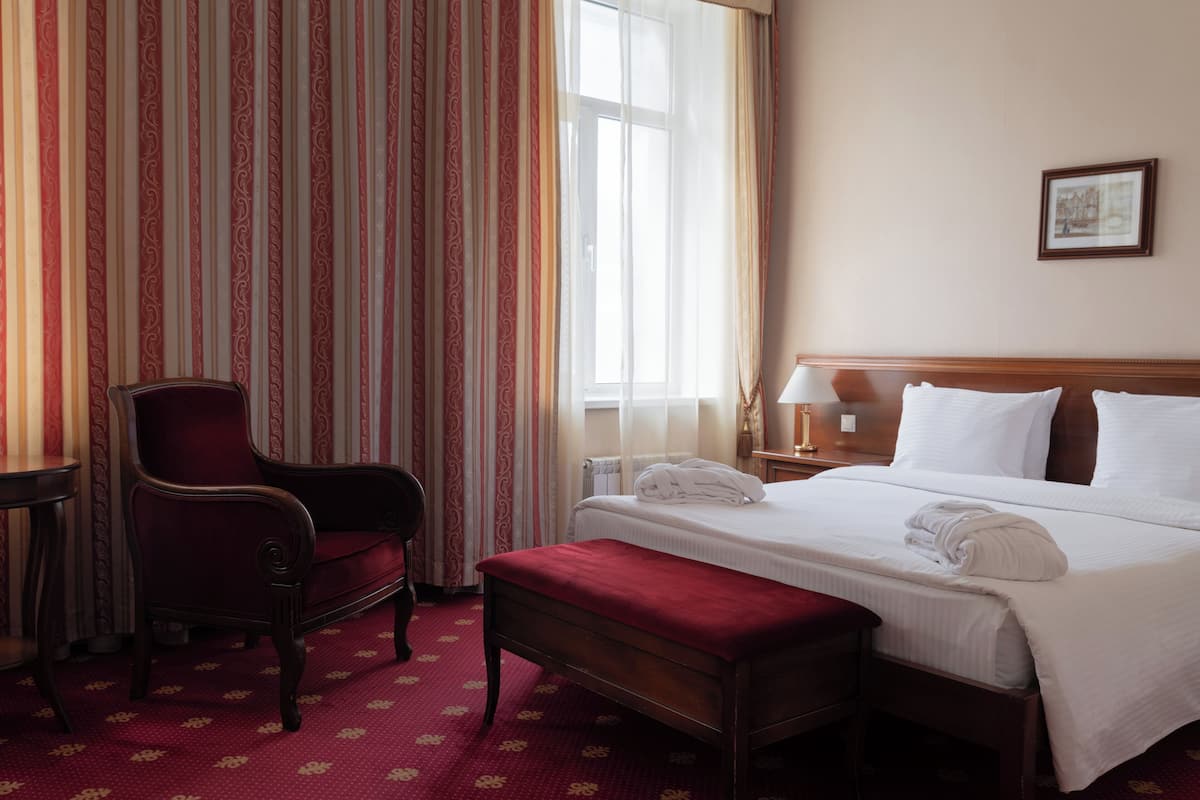 A hotel room with a bed, a chair, a bench, a nightstand, a lamp, and a window with curtains.