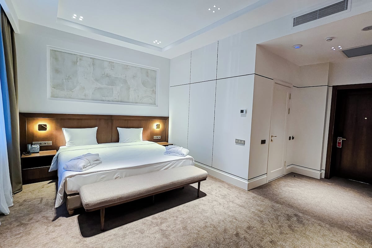A modern hotel room with a large bed, a bench, a wardrobe, and a door.
