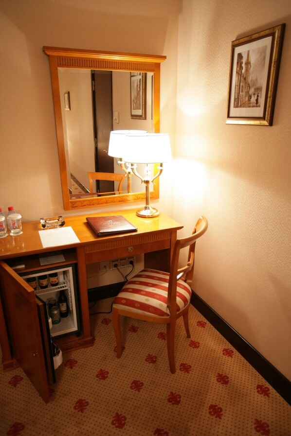 A hotel room with a wooden desk, a chair, a mirror, a lamp, a refrigerator, and a framed picture on the wall. - Grand Hotel Tien Shan (Almaty)