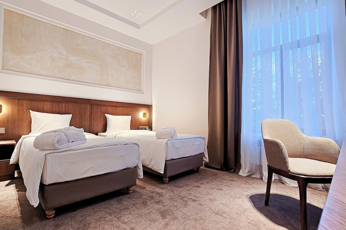 A hotel room with two beds, a large artwork on the wall, a nightstand with a lamp, and a chair by the window.