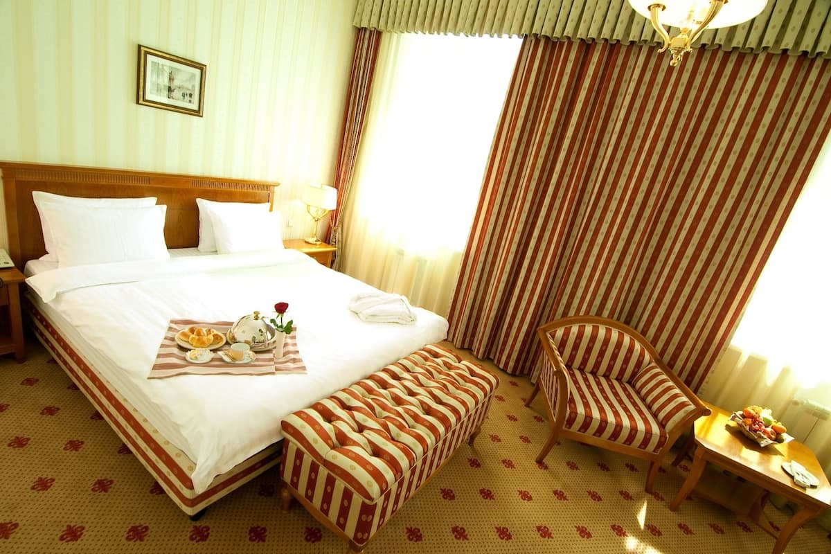 A hotel room with a large bed, a bedside table with a lamp, a chair, a small table with fruit, and a window with curtains.