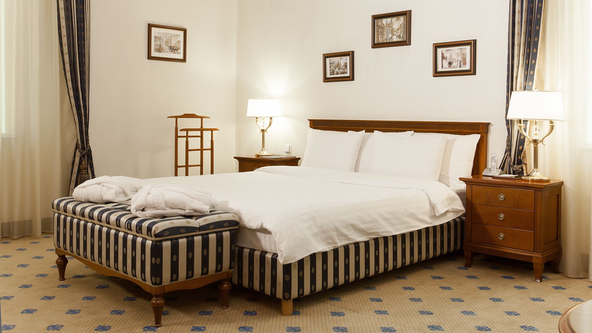 A hotel room with a large bed, a bedside table with a lamp, a small bedside table with a lamp, a wooden bench, and a patterned carpet.