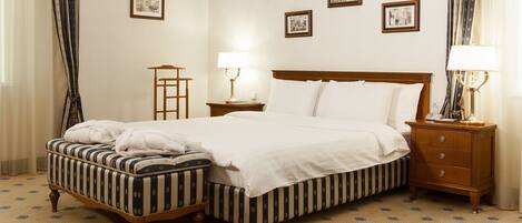 A hotel room with a large bed, a bedside table with a lamp, a small bedside table with a lamp, a wooden bench, and a patterned carpet.