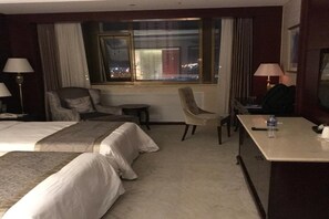 A hotel room with two beds, a desk, a chair, and a window with a city view. - Howard Johnson by Domando Plaza Wulanchabu (Ulaan Chab)