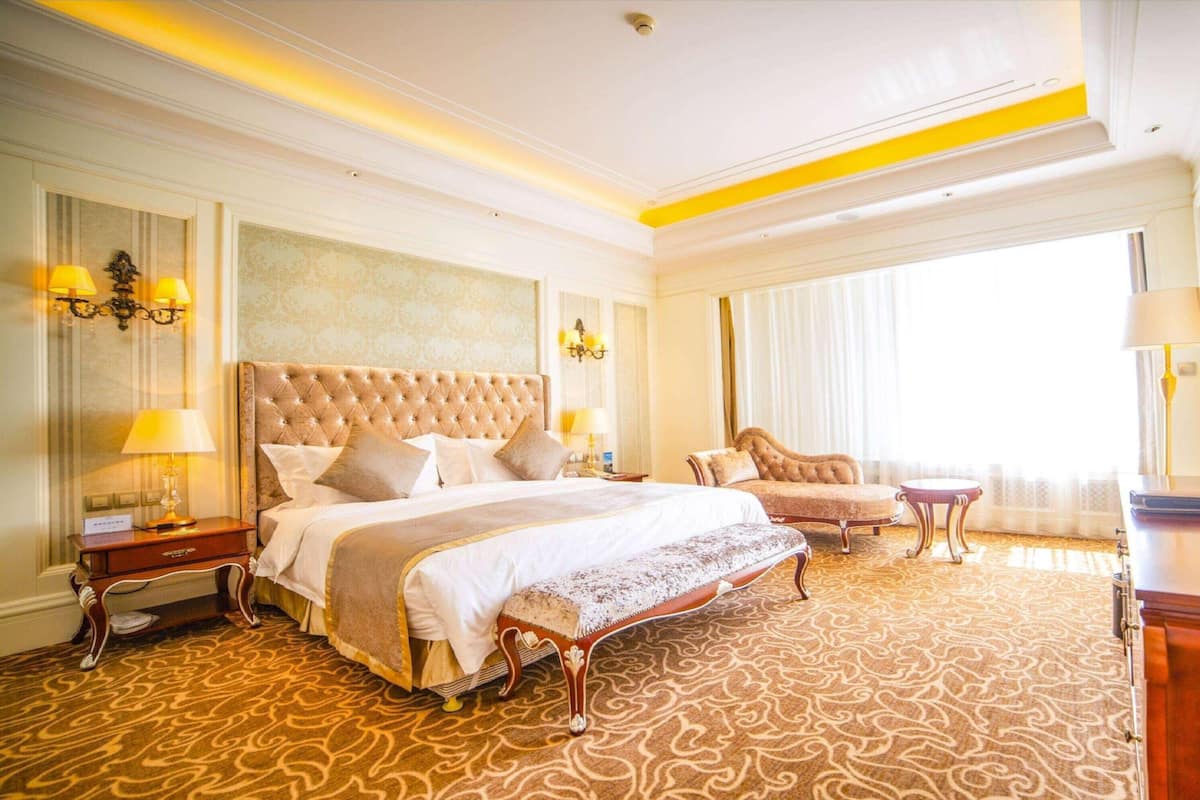 Habitación de hotel con una cama grande, un rincón de descanso con una silla, una mesita y un ventanal amplio.