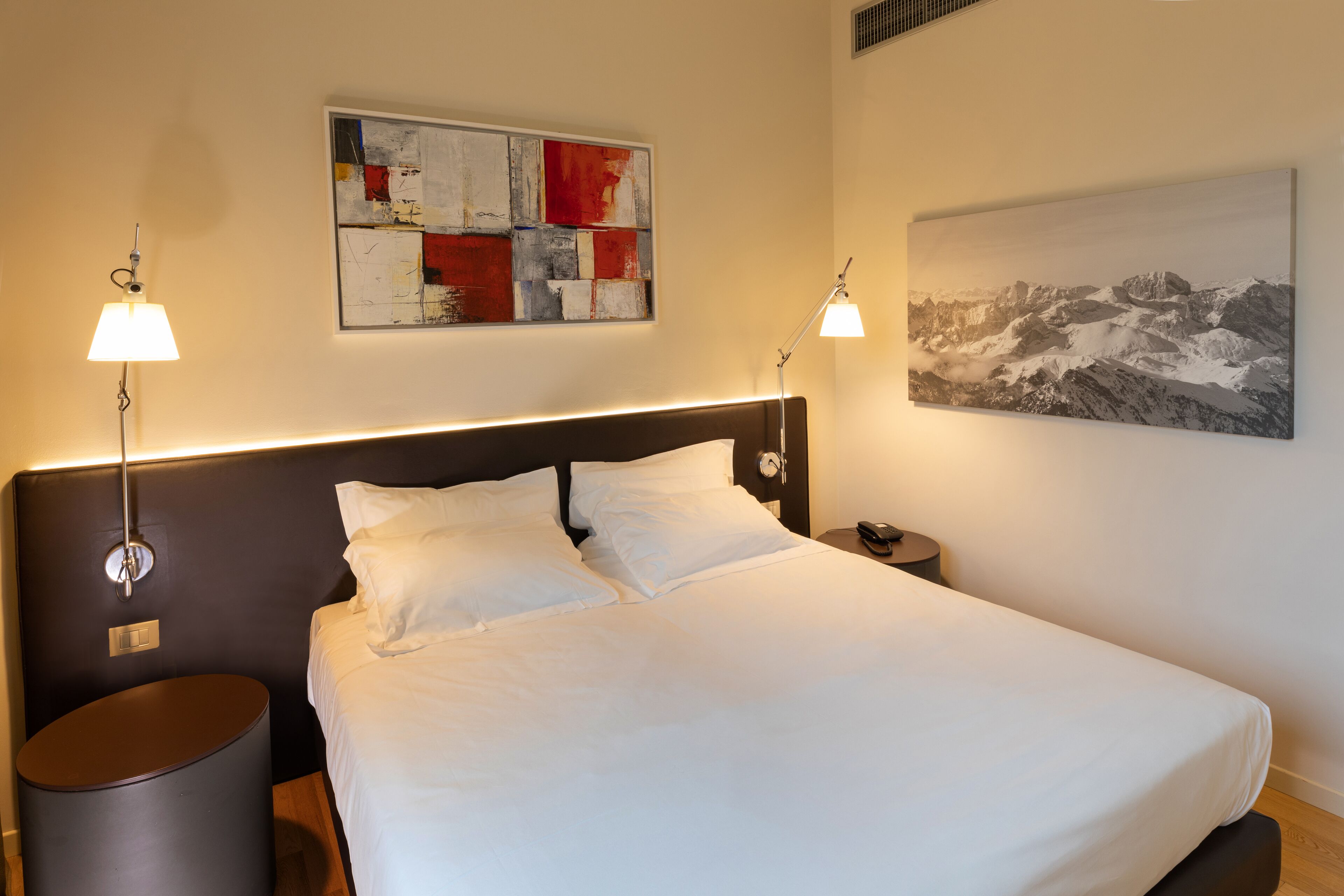 A neatly made bed with white linens, two bedside lamps, a modern headboard, and two artworks on the walls.