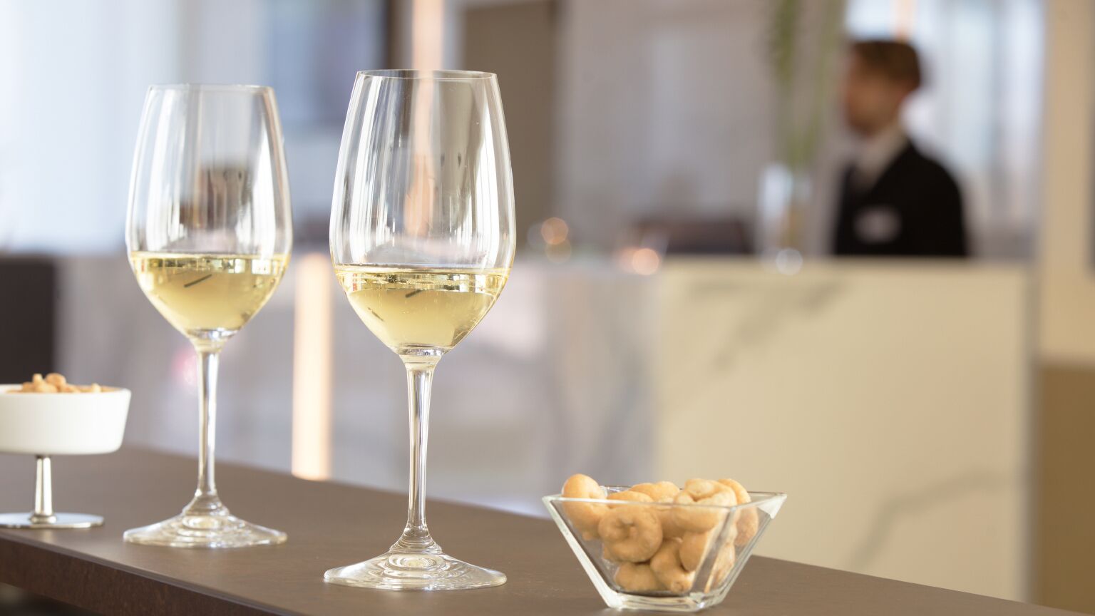 Dos copas de vino sobre una mesa, con un cuenco de frutos secos y un postre al fondo.