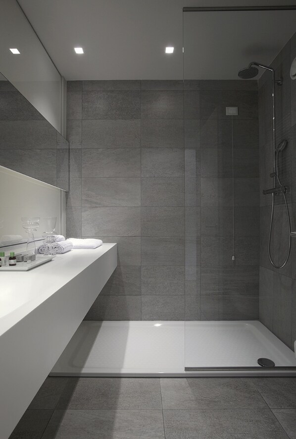 Un baño moderno con cabina de ducha de vidrio, tocador blanco con artículos de aseo y paredes con azulejos grises.