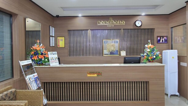 A reception desk with a wooden finish, a clock, framed certificates, and floral arrangements. - La Sara Vista (Bengaluru)
