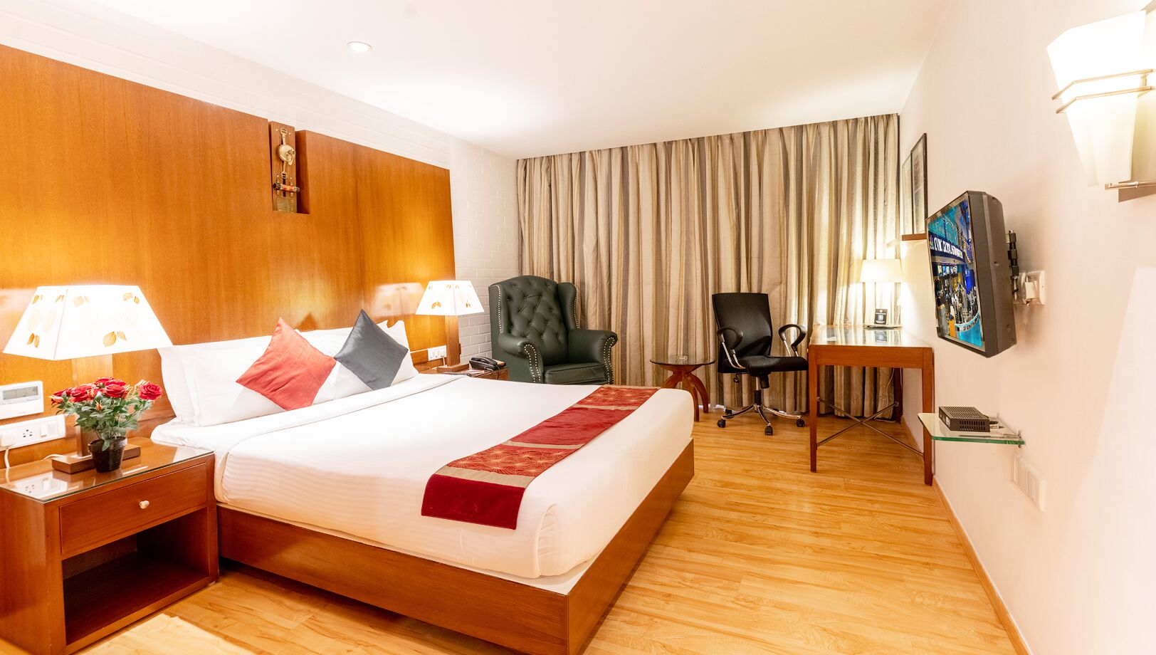 A hotel room with a large bed, two bedside tables with lamps, a green armchair, a desk with a chair, and a television on the wall.