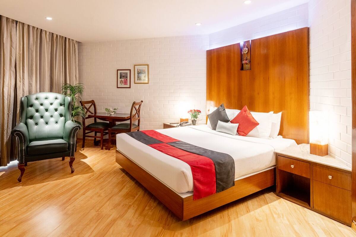 A hotel room with a wooden bed, a green armchair, a wooden nightstand, and a small table with chairs.