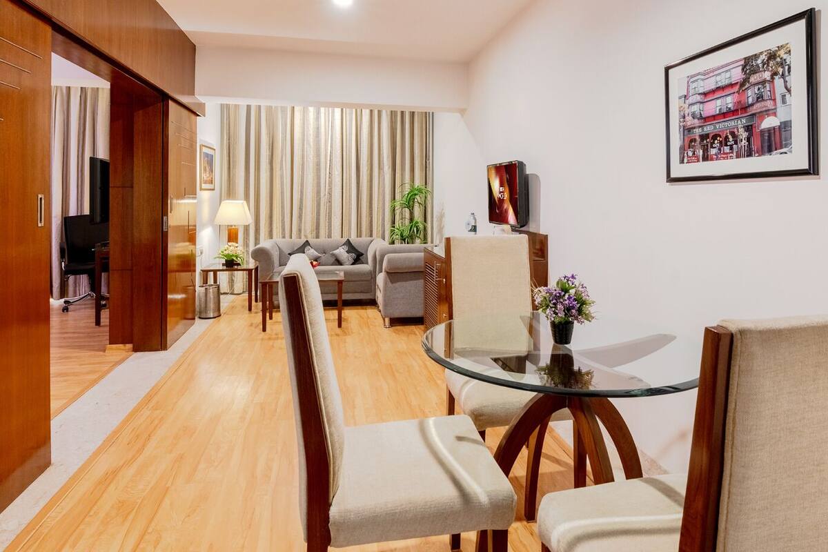 A modern hotel room with a wooden floor, a glass dining table, a sofa, and a TV.