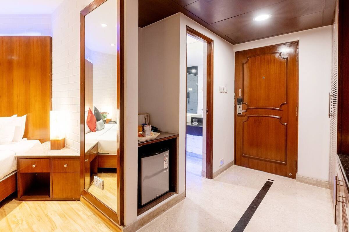 A hotel room with a bed, bedside table, mirror, wardrobe, and a small kitchenette area.