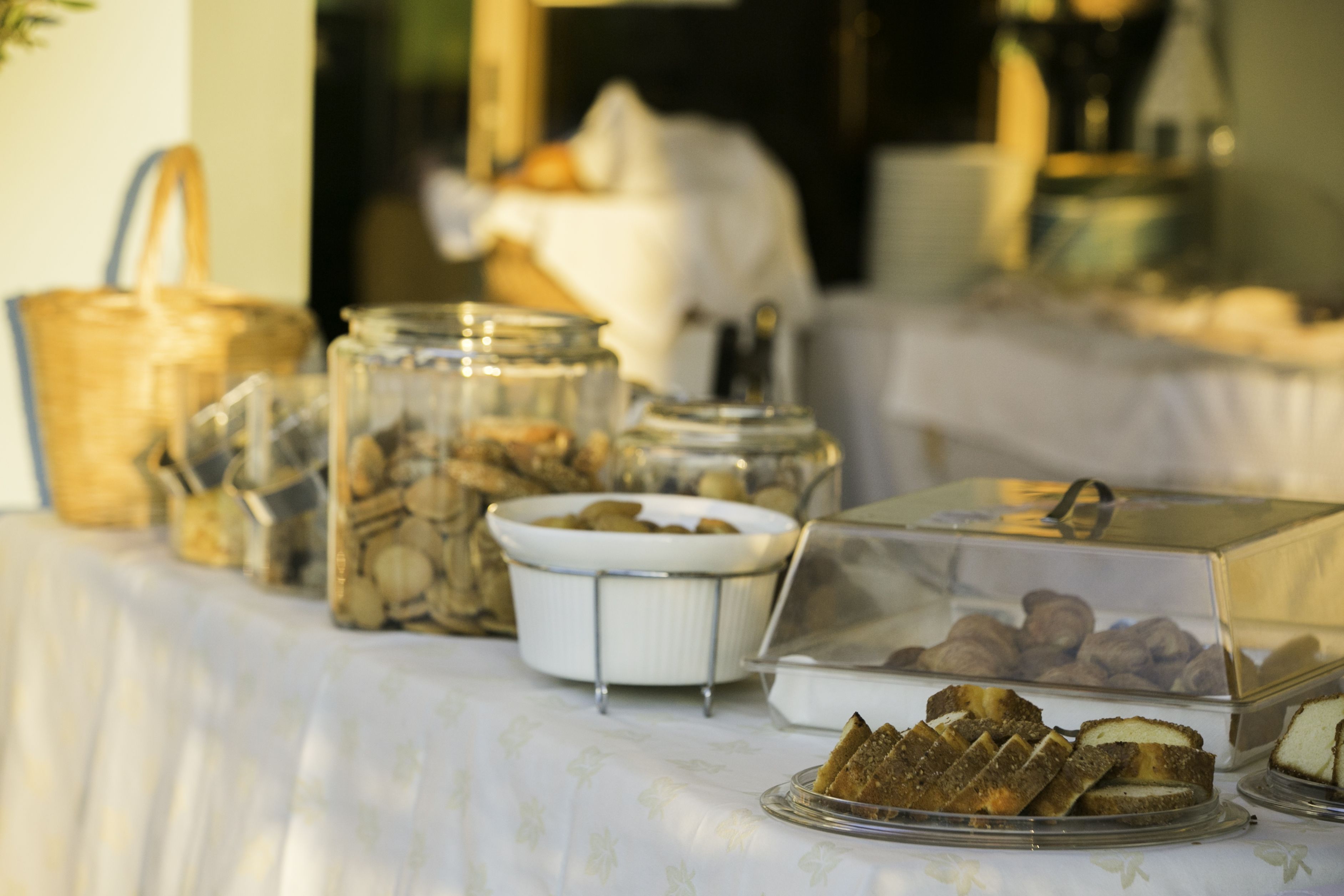 Une table de buffet avec divers plats présentés dans des récipients transparents, une nappe blanche et un sac à main en arrière-plan.