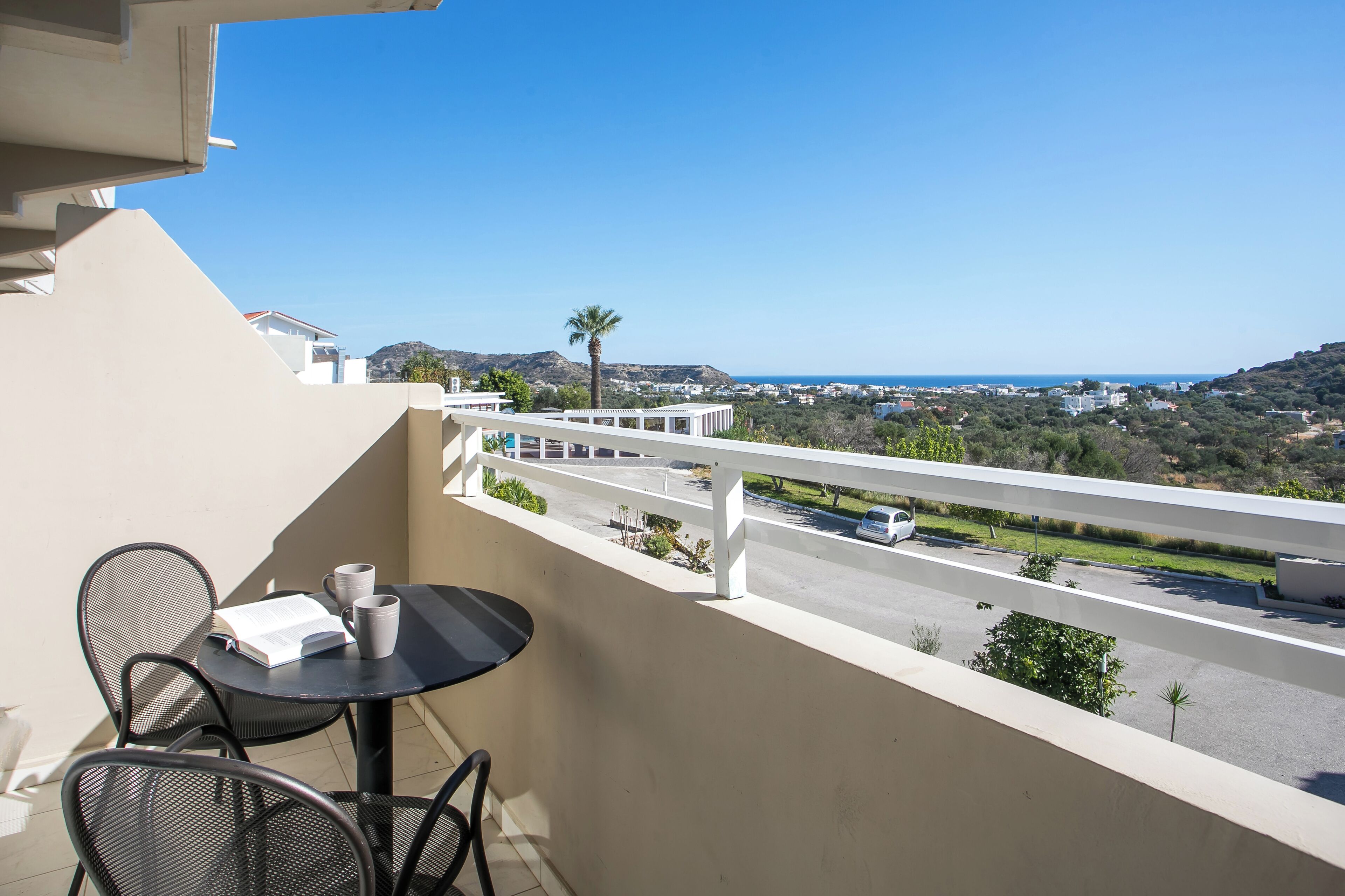 Un balcon avec une table et des chaises, donnant sur une route et sur un espace verdoyant.