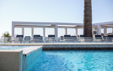 A swimming pool with clear blue water, lounge chairs, and a white pergola structure in the background.