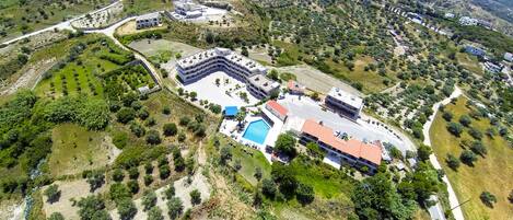 An aerial view of a resort with a large pool, multiple buildings, and surrounding greenery.