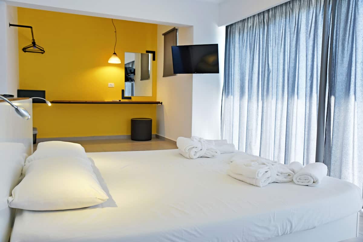 A hotel room with a bed, white pillows, a flat-screen TV mounted on the wall, a small trash bin, and a window with curtains.