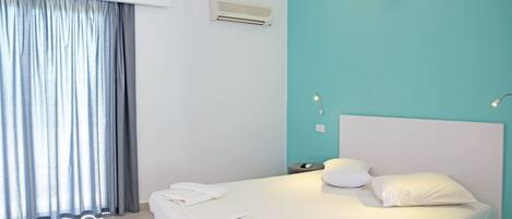 A hotel room with a bed, a window with curtains, an air conditioning unit, and a wall-mounted light fixture.