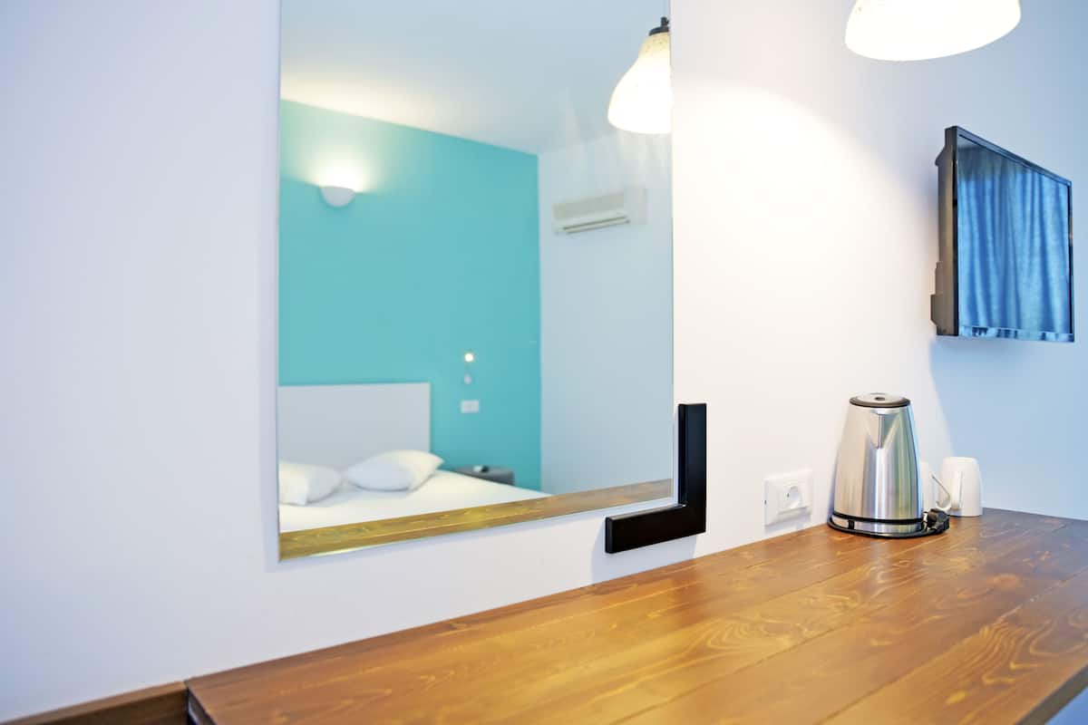 A hotel room with a wooden floor, a bed with white bedding, a wall-mounted mirror, a coffee maker, and a blue curtain.