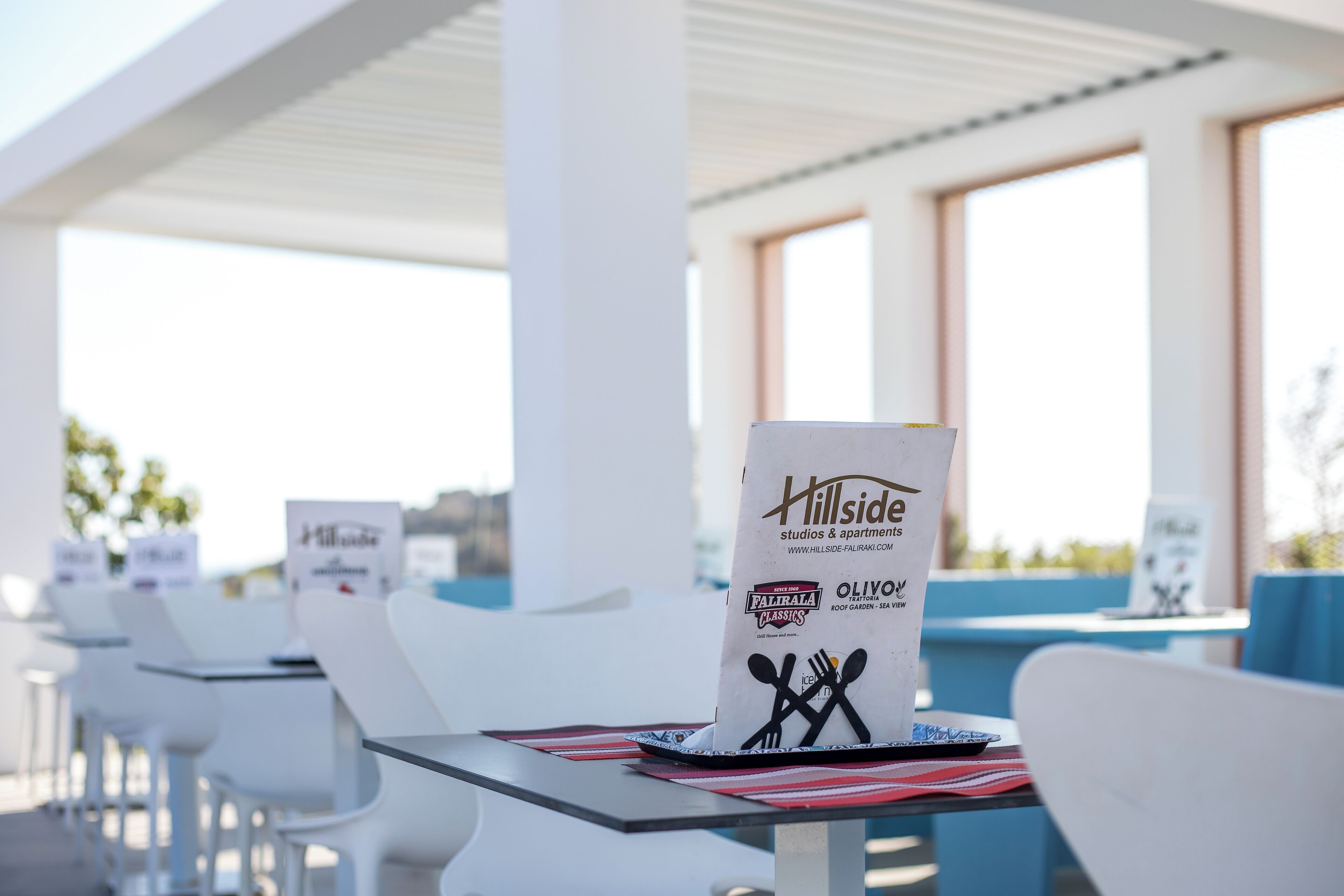 Un patio de restaurante con sillas y mesas blancas, un letrero de Hillside y un atril para el menú.