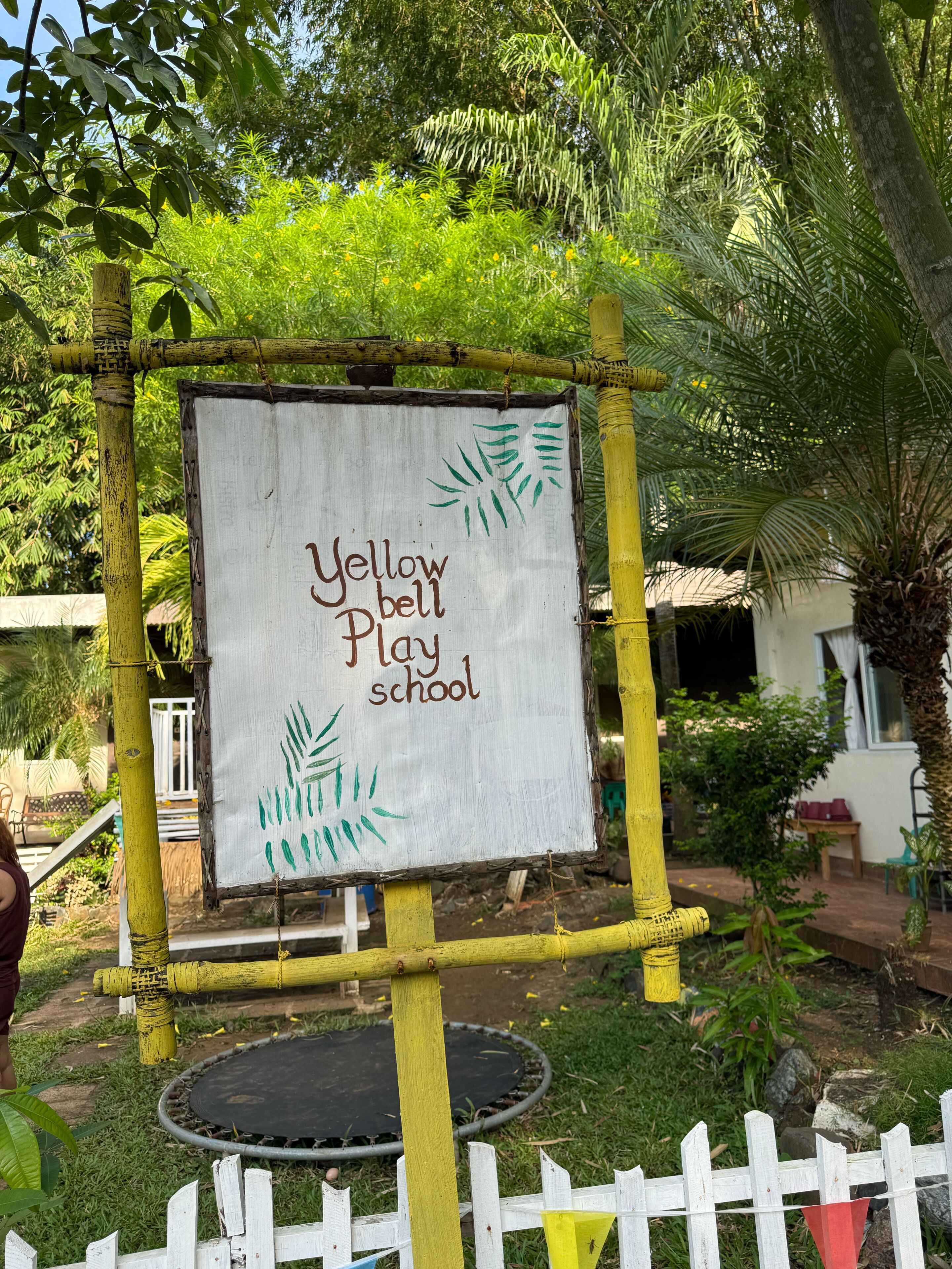 A sign for Yellow Bell Play School surrounded by greenery and a white fence.
