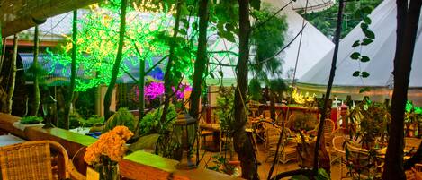 Un espace de restauration en plein air, avec des tables et des chaises, entouré de verdure et abrité par une structure en forme de tente blanche.