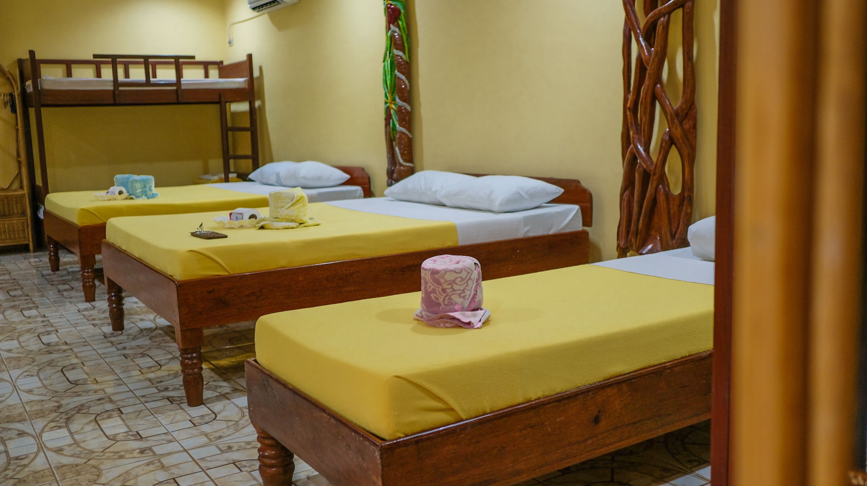 A room with three wooden beds, each with a yellow bedspread and white pillows. There is a wooden ladder against the wall, and a decorative plant with green leaves and a yellow flower.