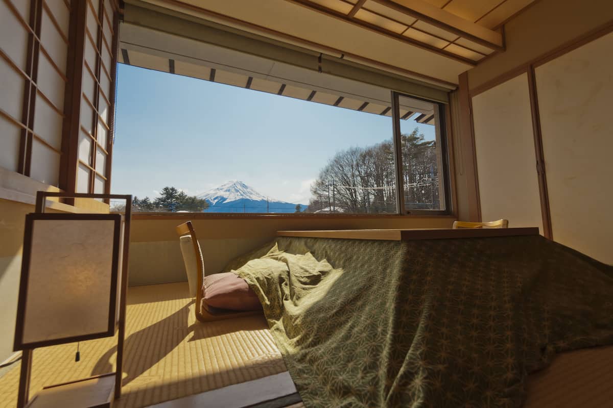 A room with a tatami mat, a futon bed, a futon chair, and a large window offering a view of a mountain.
