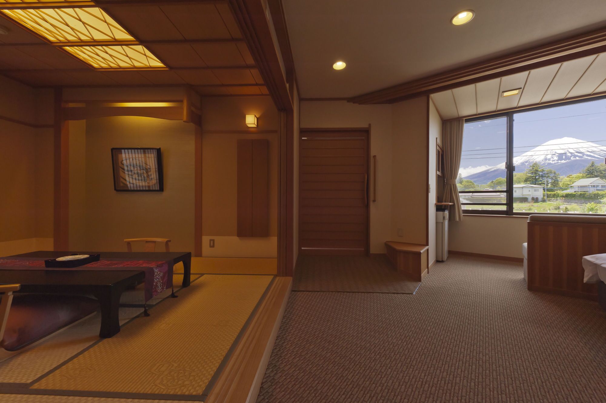 A traditional Japanese room with tatami flooring, a low table with a cushion, a sliding door, and a view of a mountain through a large window.