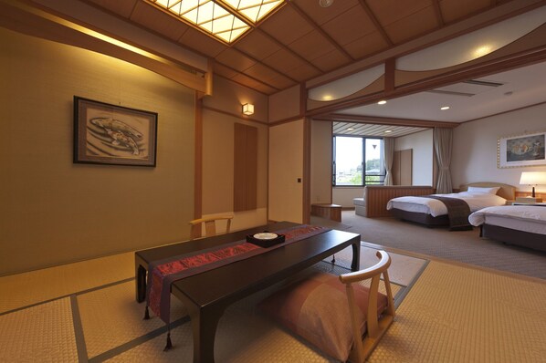 A hotel room with a bed, a desk, and a chair. There is a window with curtains, a painting on the wall, and a lamp on the bedside table.