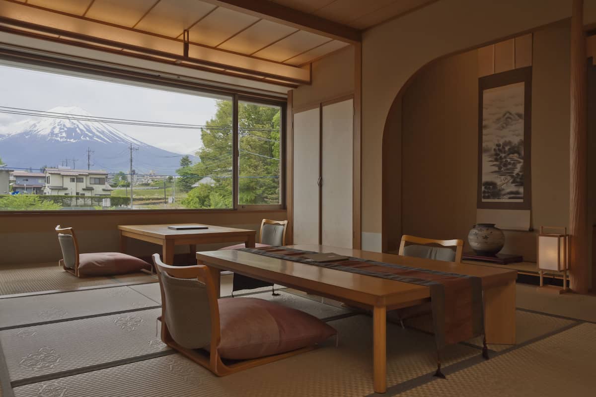 A traditional Japanese room with tatami flooring, a low table, and chairs. A large window offers a view of a mountain.