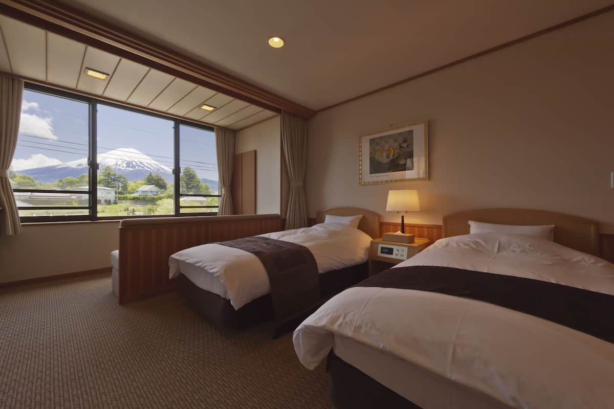 A hotel room with two beds, a window with a view of a mountain, a painting on the wall, and a bedside table with a lamp.