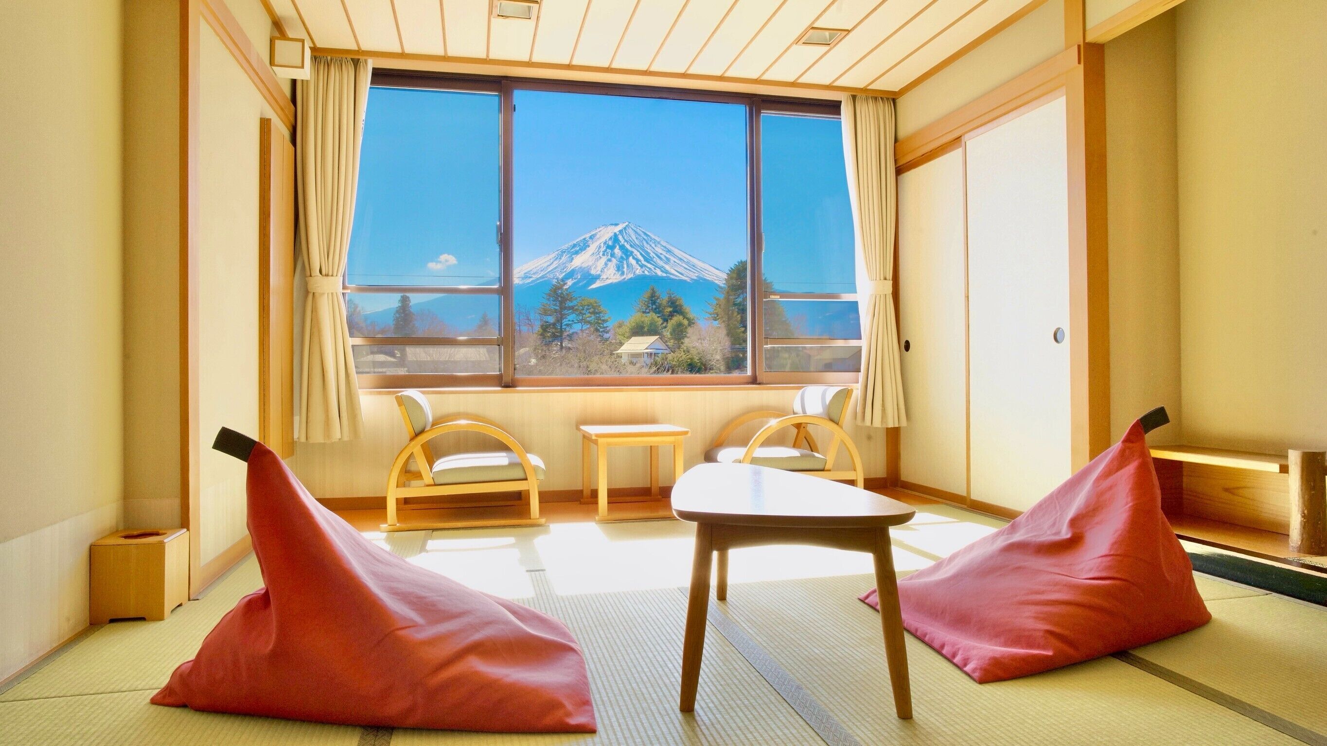 A room with tatami flooring, a low table, two chairs, and a view of a mountain.