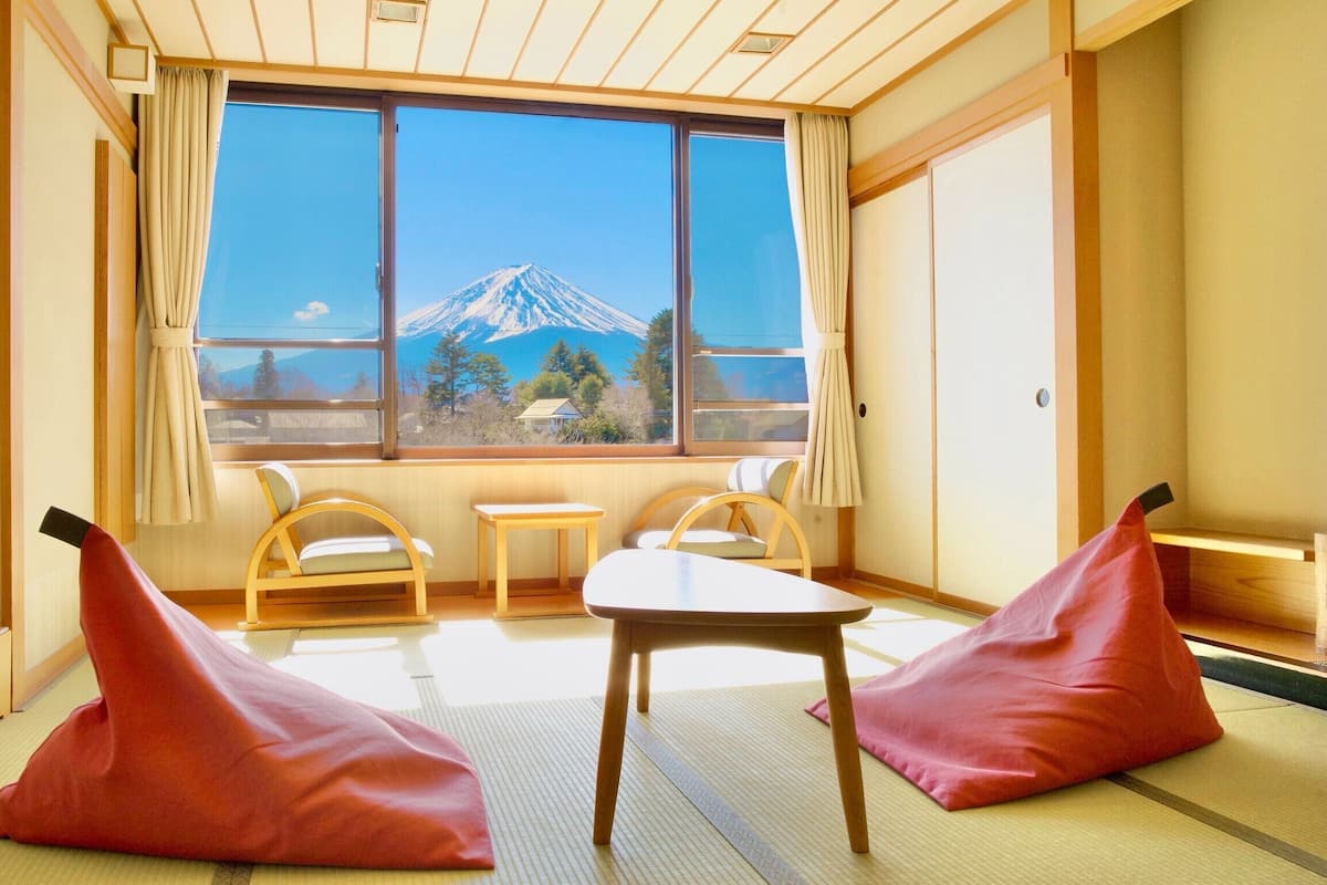 A room with tatami flooring, a low table, two chairs, and a view of a mountain.