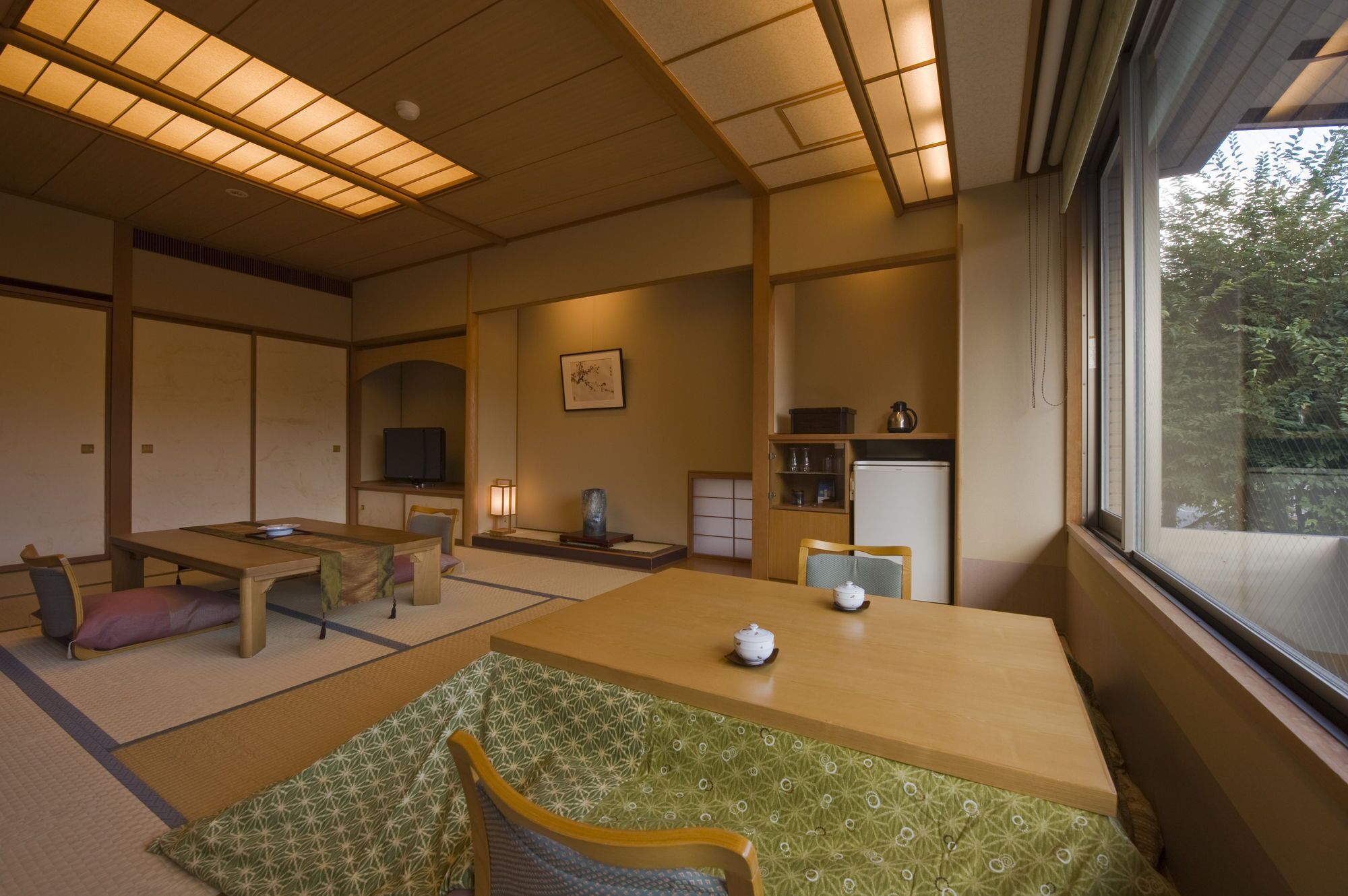 Una habitación tradicional japonesa con suelo de tatami, una mesa baja con un juego de té, un televisor y vistas a zonas verdes.