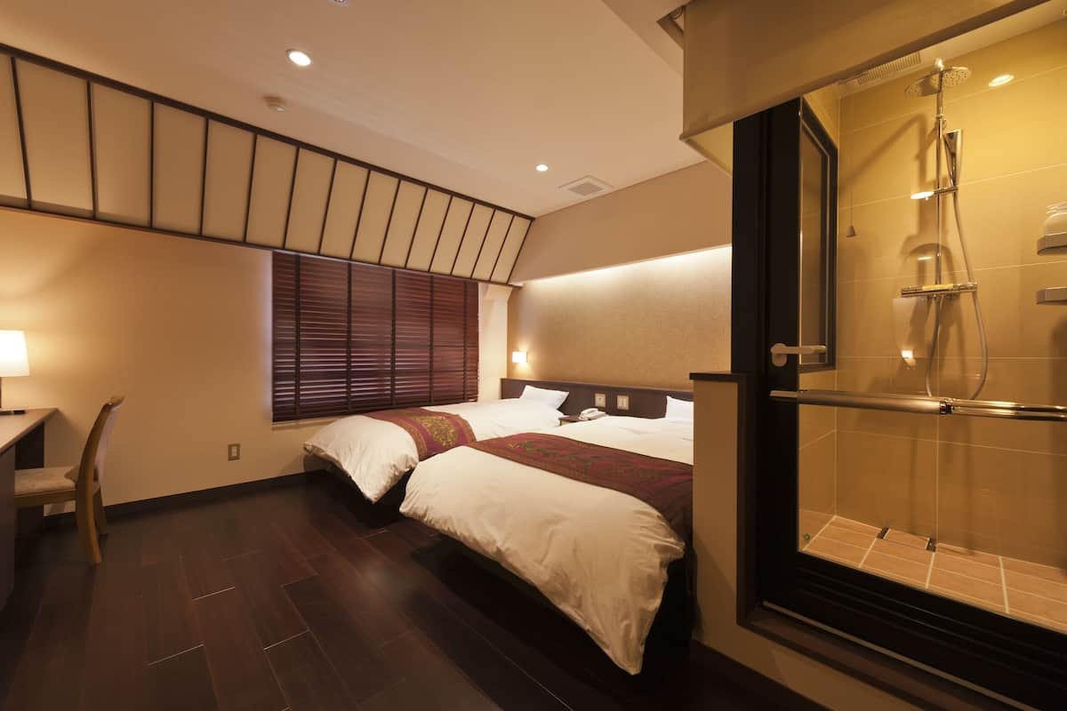 A hotel room with two beds, a desk, a chair, a shower area, and a window with blinds.