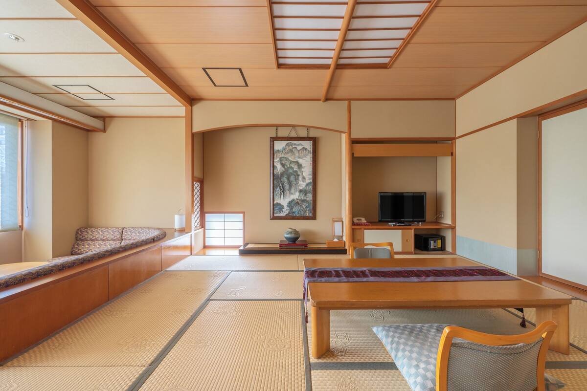 A traditional Japanese room with tatami flooring, a low table, a sofa, a television, and a painting on the wall.