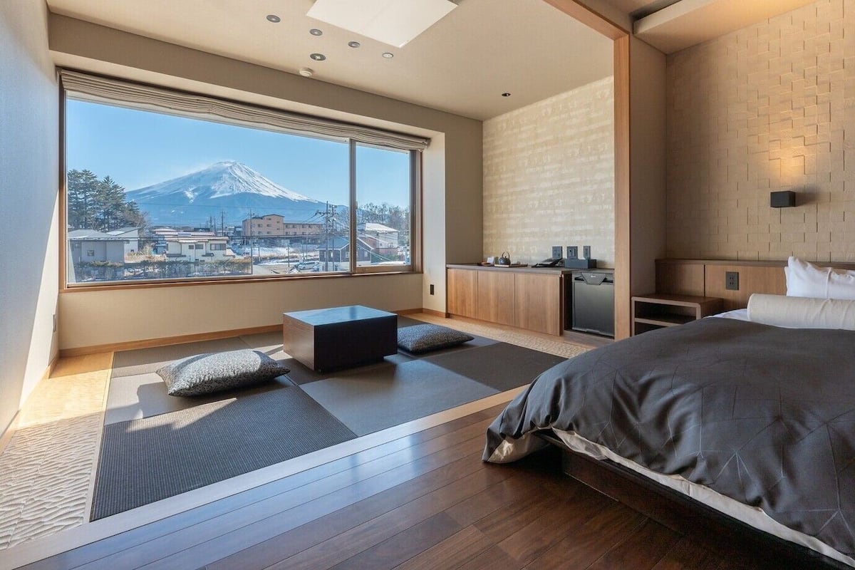 A modern hotel room with a large bed, a small seating area, a kitchenette, and a view of a mountain.