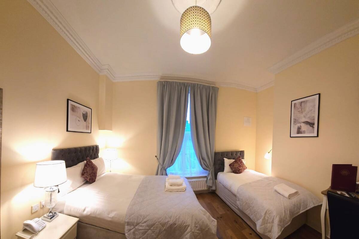A hotel room with two beds, a bedside table with a lamp, a telephone, and a window with curtains.