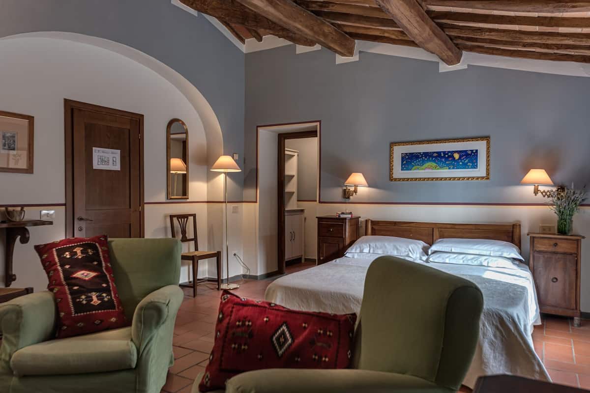 A bedroom with a bed, two chairs, a nightstand, a lamp, a painting, and a wooden ceiling.