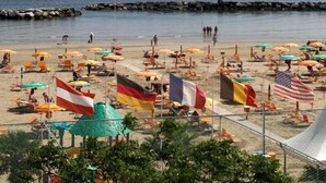 Una playa con numerosas sombrillas y banderas, un rompeolas rocoso y una vista despejada del mar.