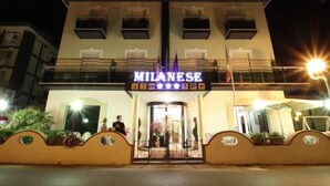 Un hotel llamado MILANESE con una entrada destacada, un camino de acceso con puerta de rejas y una fachada bien iluminada.