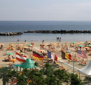 A beach with numerous umbrellas and flags, a rocky breakwater, and a clear view of the sea.