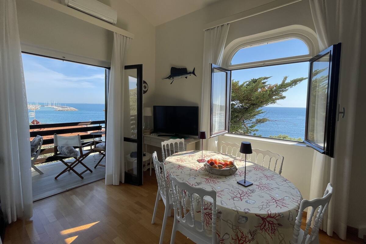 A room with a dining table, chairs, a television, and a view of the sea.