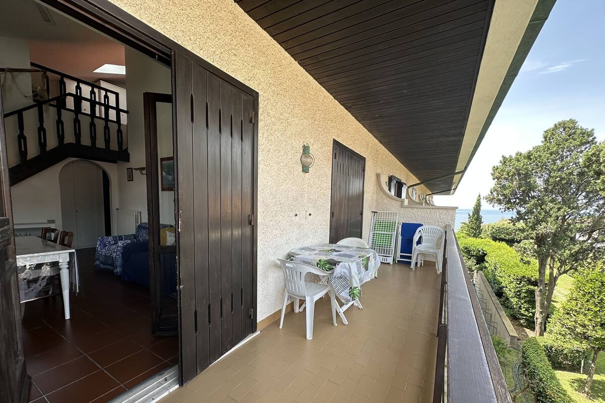 A covered outdoor area with a dining table and chairs, a staircase leading to an upper level, and a view of the sea.