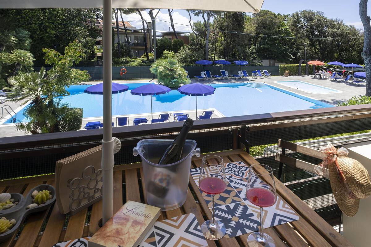 A poolside view with lounge chairs, umbrellas, and a table with a bottle, glass, and book.