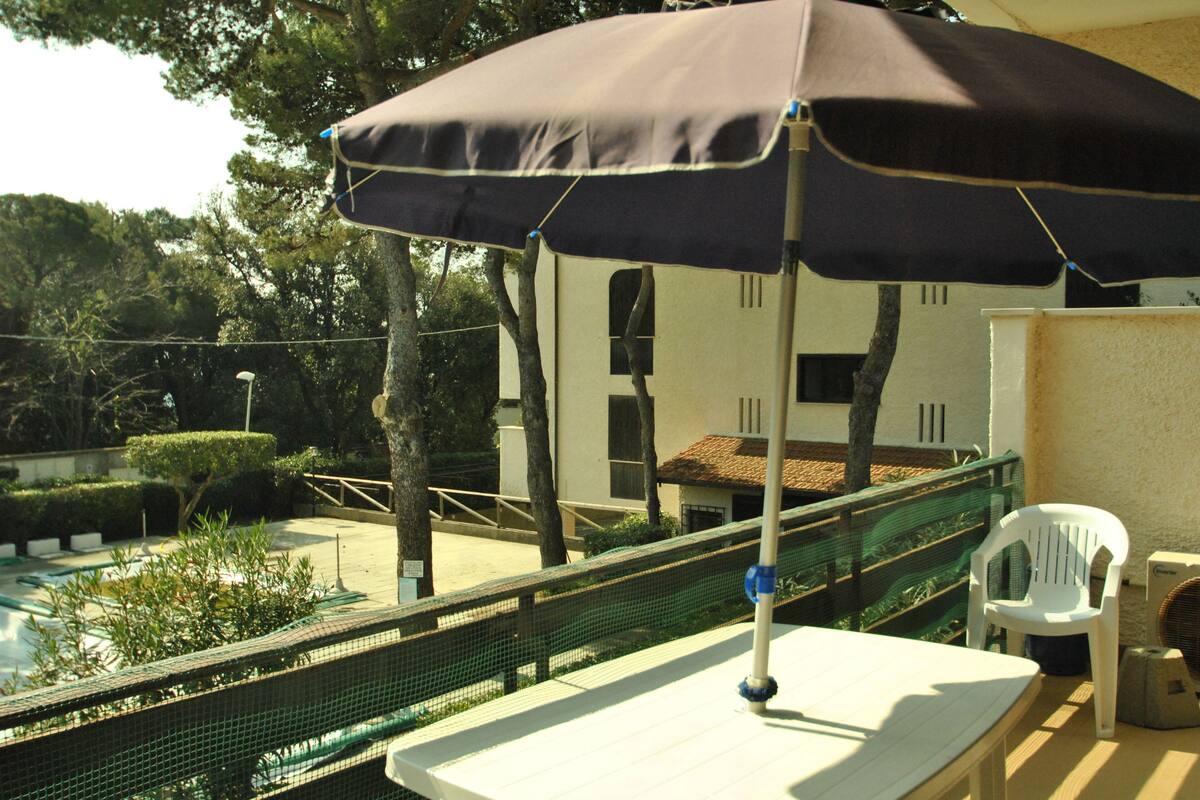 A poolside area with a table and umbrella, surrounded by greenery and a building with a tiled roof.